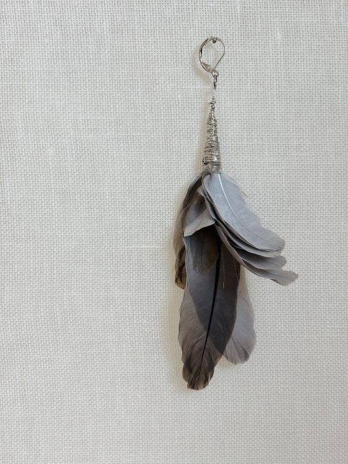 A handmade earring crafted from the organic residue of a Eurasian collared dove. A cluster of grey and brownish feathers is tightly bound at the base with silver wire, incorporating a simple metal earring hook. The composition retains the natural, slightly irregular arrangement of the feathers as they were found. The object is presented against a neutral, textured off-white fabric background, emphasizing its character as a piece of organic jewelry.