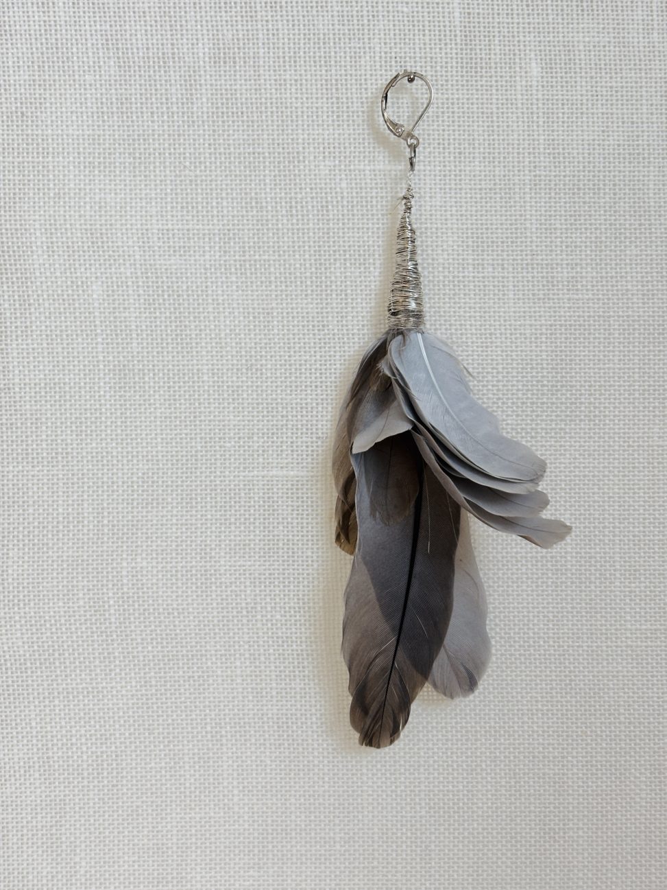 A handmade earring crafted from the organic residue of a Eurasian collared dove. A cluster of grey and brownish feathers is tightly bound at the base with silver wire, incorporating a simple metal earring hook. The composition retains the natural, slightly irregular arrangement of the feathers as they were found. The object is presented against a neutral, textured off-white fabric background, emphasizing its character as a piece of organic jewelry.